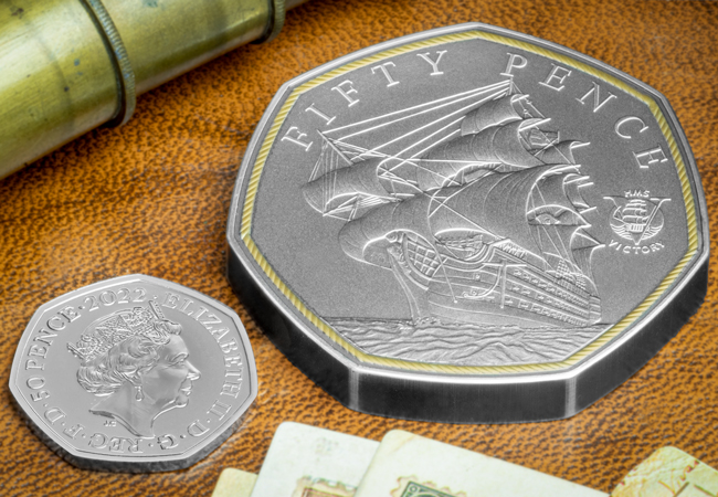 Image shows the Masterpiece HMS Victory coin next to a standard 50p coin, showing it is twice the size. The coins are lying on top of a leather-look table.